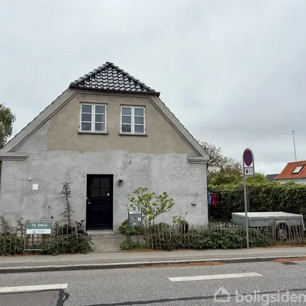Et gråt to-etagers hus med sort tag ses på et fortov ved en vej. En hvid skilt siger "TIL SALG". Hække og træer omkranser området.