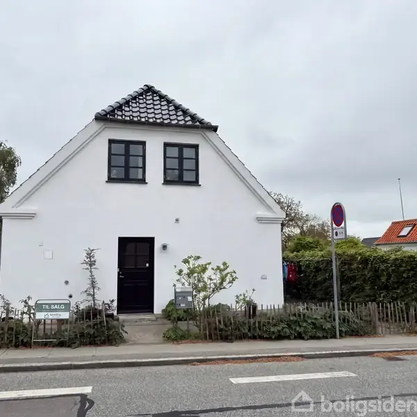 Hvidt hus med sort tag og to sorte vinduer øverst. Foran er en sti med buske og et skilt med teksten "TIL SALG". Omkring ses andre huse og en vej med fortov.