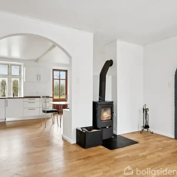 En brændeovn står i en lys stue med hvide vægge og trægulv. Bagved ses et køkken med hvide skabe og store vinduer. To buede åbninger forbinder rummene.