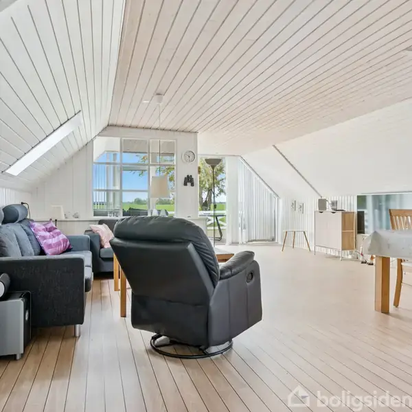 En lys stue med skråloft og trægulv. Sofa og lænestol står omkring et sofabord, mens et spisebord med stole er i baggrunden. Vinduer giver udsigt til natur.