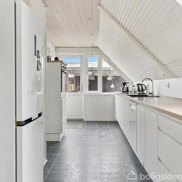 Et hvidt, moderne køkken med skrånende loft og gråt gulv. Der er en opvaskemaskine, kogeplader og køleskab. Store vinduer i baggrunden giver lys. En kop står på bordet.