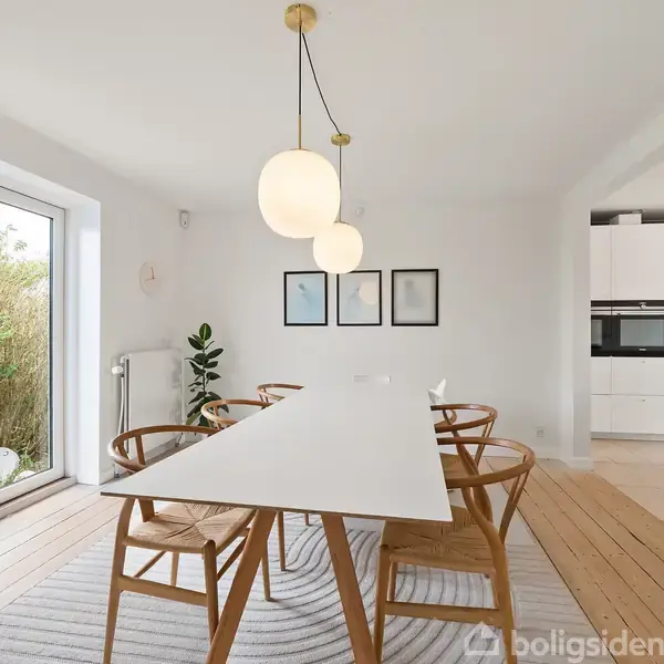 Et rektangulært spisebord med seks træstole i en lys spisestue. Hængende lamper over bordet og tre små vægkunstværker på væggen. Store vinduer lader naturligt lys ind.