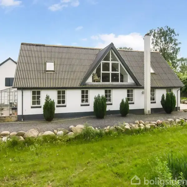 Et hvidt hus med gråt tag står i en grøn have. Huset har store vinduer og en skorsten. Foran er en stenbelægning og buske, omgivet af græs og træer.