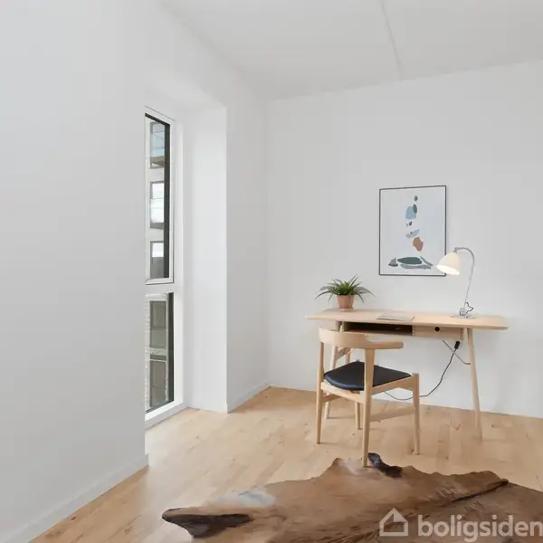 Et skrivebord står i et lyst rum, med en plante og lampe på det. Der hænger et billede over skrivebordet, og en stol står foran det. Store vinduer bringer lys ind.