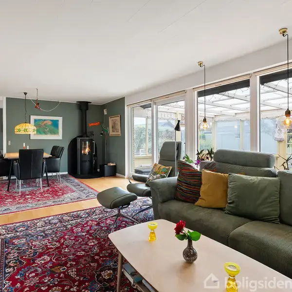Stue med grøn sofa fyldt med farverige puder foran et sofabord med små vaser. I baggrunden står en brændeovn, spisebord og tæppe. Store vinduer viser en overdækket terrasse.