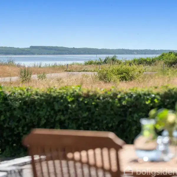 Træbord med blomster og glas står i en have foran en tæt hæk. I baggrunden ses en strand og en rolig kystlinje med grønne områder under en klar blå himmel.