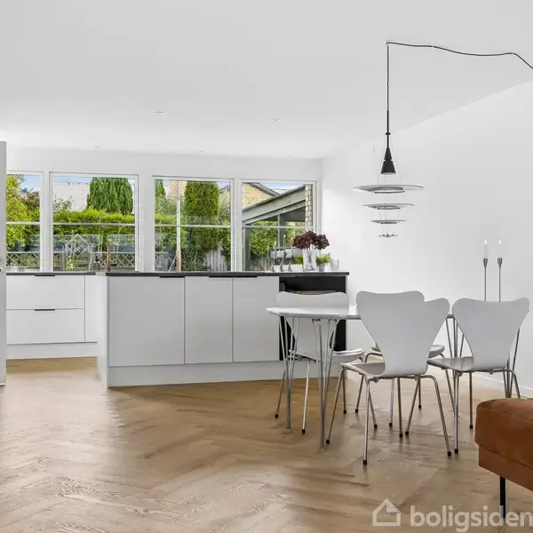 Et lyst køkken-alrum med moderne spisebord og hvide stole. I baggrunden står et hvidt køkken med store vinduer, der giver udsigt til en grøn have. En brun sofa ses til højre.