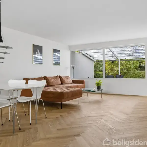 En moderne stue med en brun sofa står på et trægulv. Ved siden af er et spisebord med hvide stole. Store vinduer viser en terrasse og grøn beplantning udenfor.