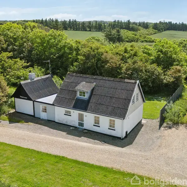 Et hvidt murstenshus med sort tag står stille omgivet af frodig grøn natur og buske. En grusvej løber langs huset, og der er marker i baggrunden.