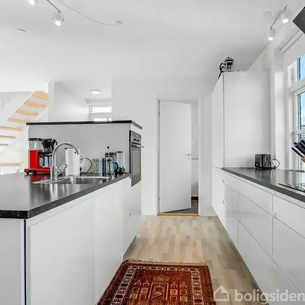 Hvidt køkken med sorte bordplader og rødt gulvtæppe. Køkkenredskaber, kaffemaskine, og olier på bordet. Trappe i baggrunden leder til første sal. Vinduer lader naturligt lys ind.