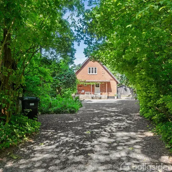 Et rødt murstenshus med et spidst tag står for enden af en grusvej, omgivet af tætte, grønne træer og buske. En terrasse med havemøbler ses foran huset.