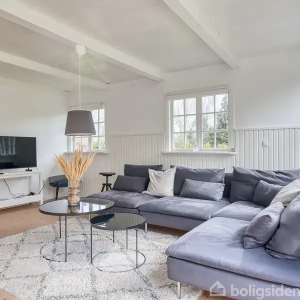 Grå sofa står i hjørnet på et lyst stuegulv. En rund sofabordsgruppe med dekoration foran sofaen. Fjernsynet står til venstre. Store vinduer giver lysindfald.