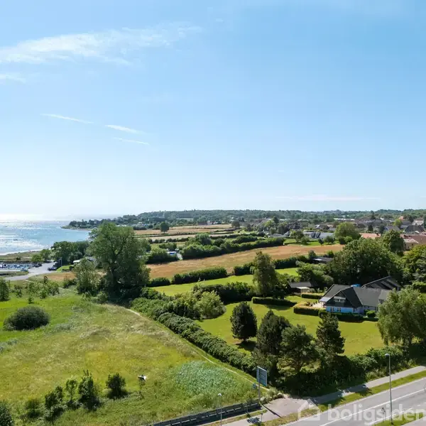 Vej strækker sig gennem frodigt landskab. Hus ligger ved vejkant omgivet af træer. Baggrund viser havn og åbent vand under klar blå himmel, skaber idyllisk kystnært miljø.