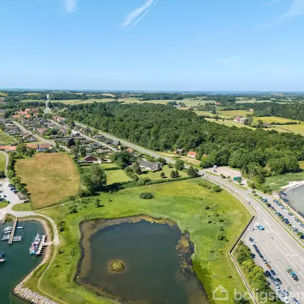 En marina med flere både fortøjet, ved siden af en vej langs en kystlinje. Omkringliggende grønne områder og skov strækker sig mod horisonten, under en klar blå himmel.