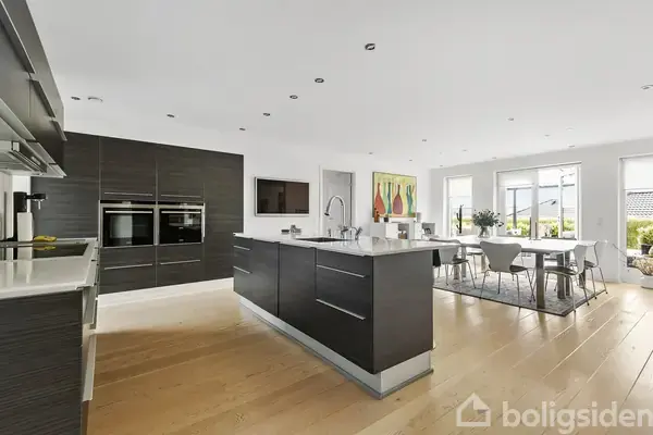 Moderne køkken i åbent rum med køkkenø i træ og stål, indbyggede apparater og spiseplads. Store vinduer giver naturligt lys, og farverig kunst pryder væggen. Lyst trægulv dominerer.