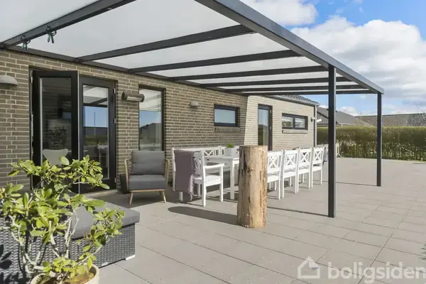 Terrasse med et langt spisebord og stole står under en overdækning. Indrammet af murstensvægge og vinduer, er der udsyn til en stor flisebelagt gårdhave med planter i potter.