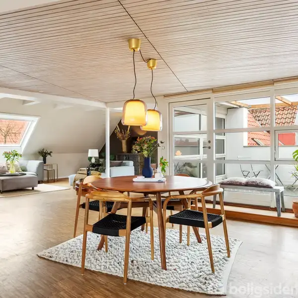 Spisebord med fire stole står centralt i et lyst rum med trægulv. Omkring ses en stue med sofa, vinduer og masser af naturligt lys. Planter står i hjørnet.