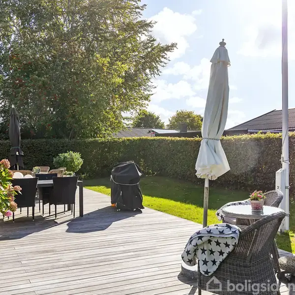 Terrasse med træbord og stole står under en paraply ved siden af en havegrill. Omkranset af buskads og træer, ses et hyggeligt udeområde i solskin.
