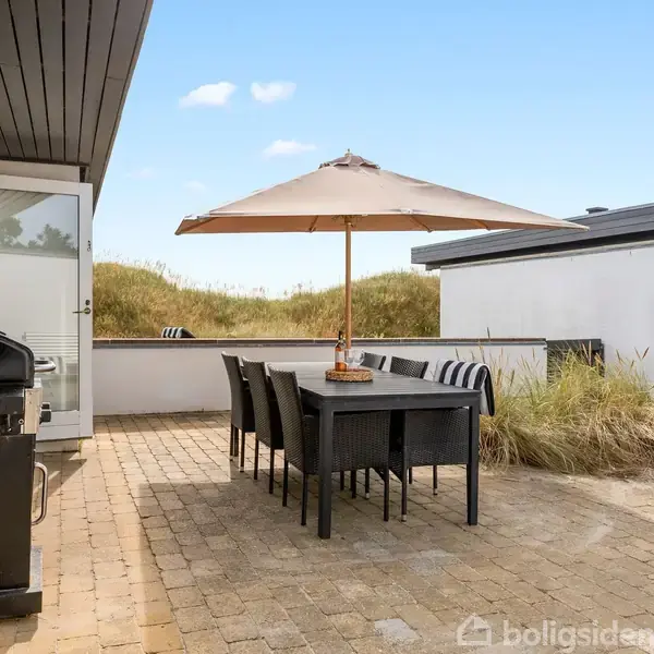 En terrasse med et udendørs spisebord og stole under en parasol. Ved siden af står en grill. Omgivelserne inkluderer sand og planter, der giver en strandagtig atmosfære.