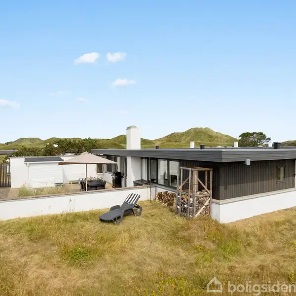 En moderne bungalow med sorte og hvide elementer står på en græsplæne. To liggestole er placeret udenfor. Landskab med bakker og blå himmel i baggrunden.