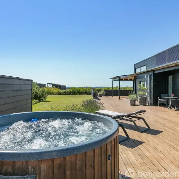 Boblebad aktivt på en solrig træterrasse ved moderne, sort-hus, omgivet af grøn have og klar, blå himmel. Lænestole placeret ved siden af til afslapning.