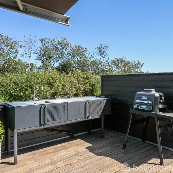 En udendørs terrasse med en sort grill står på et trædæk ved siden af en udendørs håndvask. Grønt løv i baggrunden under en blå himmel.