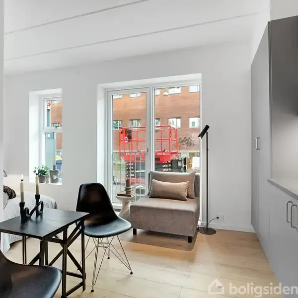 Stue med sort spisebord og stole, ved siden af en grå lænestol. Køkkenet til højre. Store vinduer med udsigt til bygning. Gulvlampe ved siden af stolen.