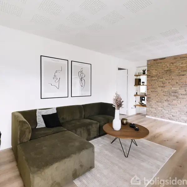 En grøn sofa står ved en hvid væg med to abstrakte billeder. En rund træbord med dekorativ vase er placeret foran. Rummet har trægulve og en halvmur af mursten.