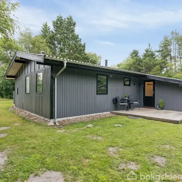 Et gråt træhus med sort tag står på en grøn græsplæne. Huset har store vinduer og en terrasse med havemøbler. Omgivelserne er skovrige med tæt bevoksning.