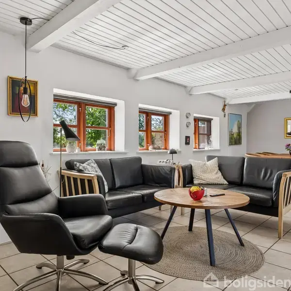Lænestol og sofa står omkring et rundt træbord med en rød skål. Stuen har hvide vægge, træloft og flere vinduer. I baggrunden ses et spisebord.