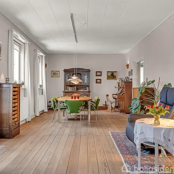 Stue med træmøbler og spisebord i midten omringet af grønne stole. En lampe hænger over bordet. Rummet har lyse vægge, store vinduer og er dekoreret med planter og kunst.