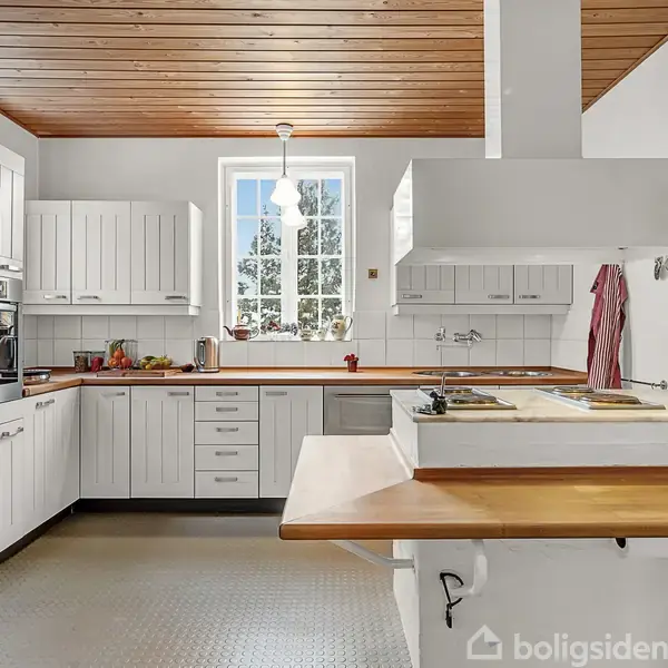 Et moderne køkken med hvide skabe og træbordplader. Køleskab og ovn i rustfrit stål til venstre. Et vindue i midten med udsigt til træer. Træloft overalt.