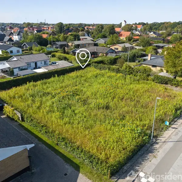 En ubebygget grund med højt græs er omgivet af huse i en villakvarter. Et digitalt stedmærke angiver placeringen. Der er en vej til højre for grunden.