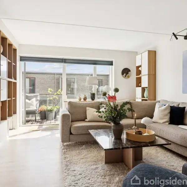 Stue med beige sofa, sofabord med vase, og bogreol langs væggen. Sollys strømmer ind fra en altan med planter. Kunst hænger på væggen. Hyggelig og moderne atmosfære.
