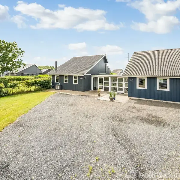 Et mørkeblåt træhus står stille i et grønt, beplantet område. Foran huset er der en grusbelagt indkørsel, og til venstre ses en velplejet græsplæne. Husnummeret "46" vises ved indgangen.