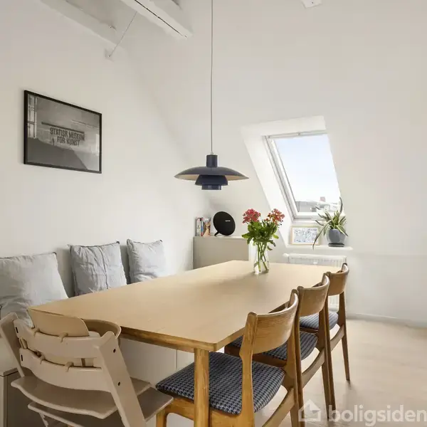 Et spisebord med stole står i et lyst rum med skråvægge. Der er puder på bænken, en hængelampe over bordet, og et vindue med udsigt.