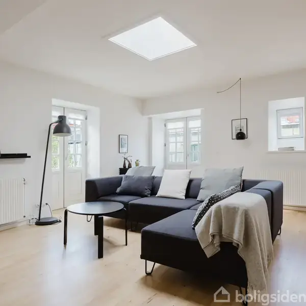 En sort hjørnesofa med tæppe og puder står i en lys stue med trægulve. Et rundt sofabord og en stående lampe er placeret ved siden af sofabordet.