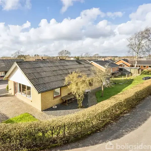 Et enfamiliehus med græsplæne og indkørsel, beliggende på en hjørnegrund omgivet af en hæk. Skyer fylder himlen, mens flere huse kan ses i baggrunden.