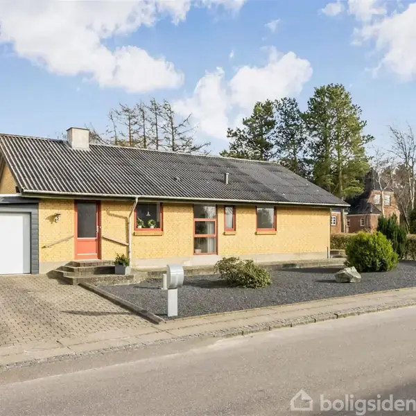 Et gult, enetages hus med sort tag står ved en vej. Der er en indkørsel med garage, grusbed og græsplæne. Træer og buske omringer huset.