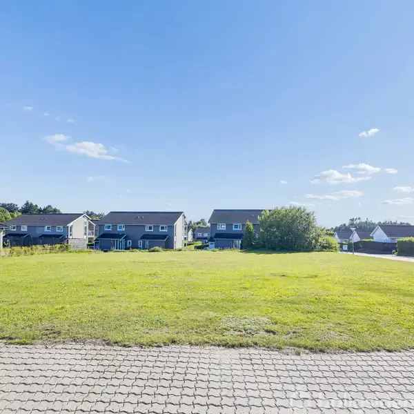 Grøn græsplæne strækker sig foran rækkehuse, der står ensartet. Husene er omgivet af en blå, skyfri himmel, med enkelte buske og et skovområde i baggrunden.