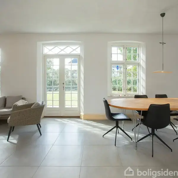En lys stue med en grå sofa til venstre og et rundt spisebord med sorte stole til højre. Store vinduer og glasdør åbner ud til en grøn have.