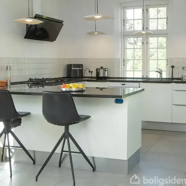 Et moderne køkken med en sort bordplade, hvorpå der står en skål med frugt. To sorte barstole er placeret ved øen. I baggrunden er der et vindue og hvide skabe.