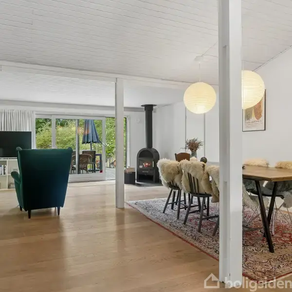 Stue med spisebord og stole, dækket af lammeskindsunderlag, på et orientalsk tæppe. Brændeovn står i hjørnet. Store vinduer med udsigt til en have i baggrunden.