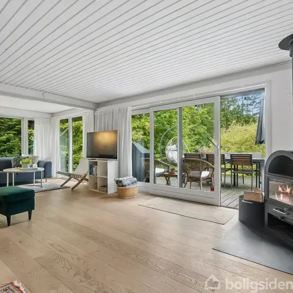 En moderne stue med en brændeovn i hjørnet, der brænder fredeligt. Rummet er lyst med store glasdøre, der åbner ud til en terrasse og en frodig have.