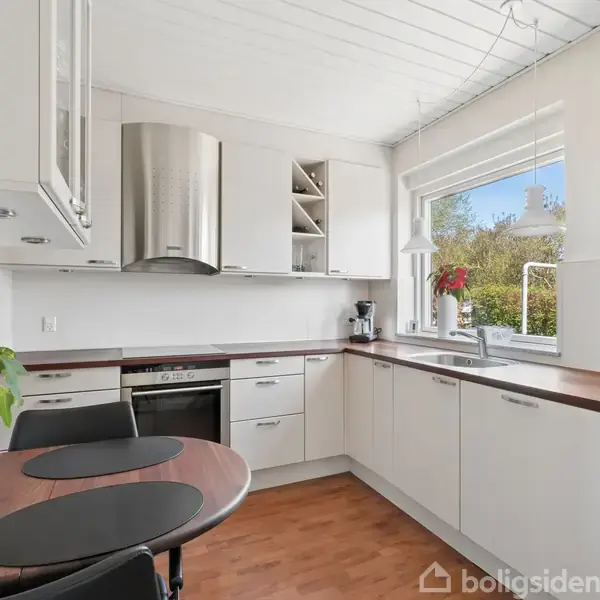 Køkkenbord med to sorte dækkeservietter, omgivet af hvide skabe og rustfri apparater. Et vindue med udsigt til grønt udenfor. Kassen lysestage og plante på bordet.