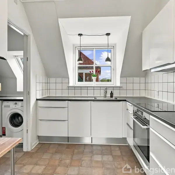 Moderne køkken med hvide skabe og sort bordplade. Et vindue med lamper over en vask giver udsigt til røde tagsten. En vaskemaskine står i det tilstødende rum.