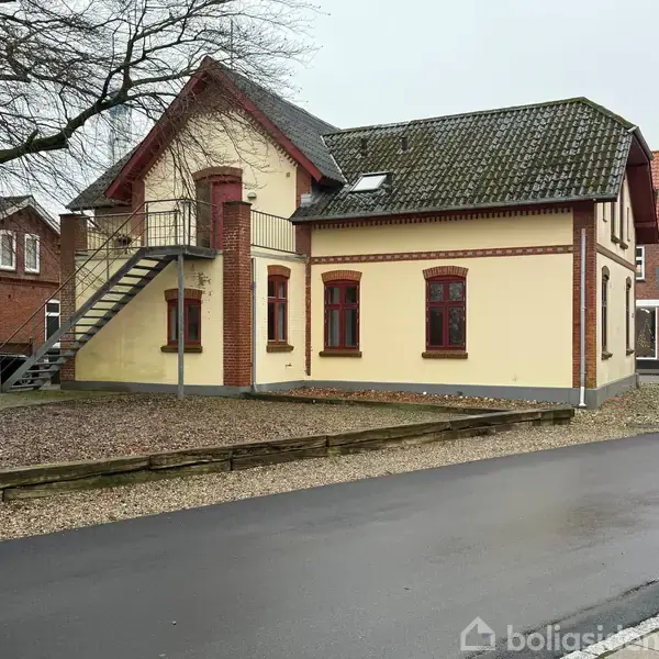 Gul murstensbygning med røde vinduesrammer, har udendørs metaltrappe. Den står ved en asfalteret vej omgivet af grus og små træhuse i baggrunden. Ingen tekst fundet.