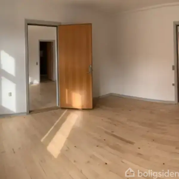 Et tomt værelse med trægulv, åbne døre, og sollys strømmer gennem vinduet. Væggene er hvide, og der er to døråbninger, der fører til andre rum.