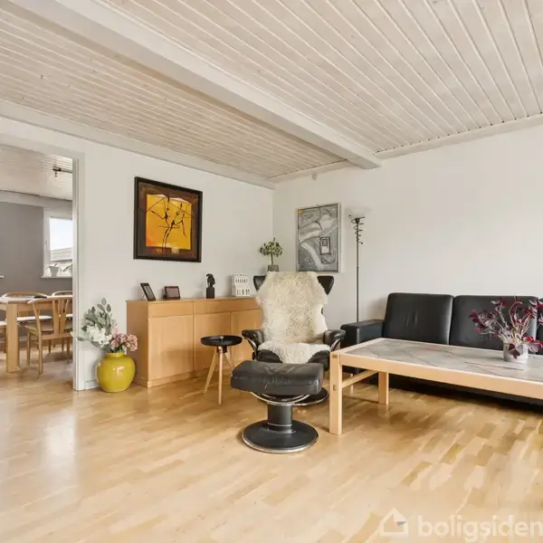 Sort sofa og stol i en moderne stue med lyst trægulv og hvide vægge. Kunst og planter pynter rummet, der åbner op til en spisestue med træmøbler.