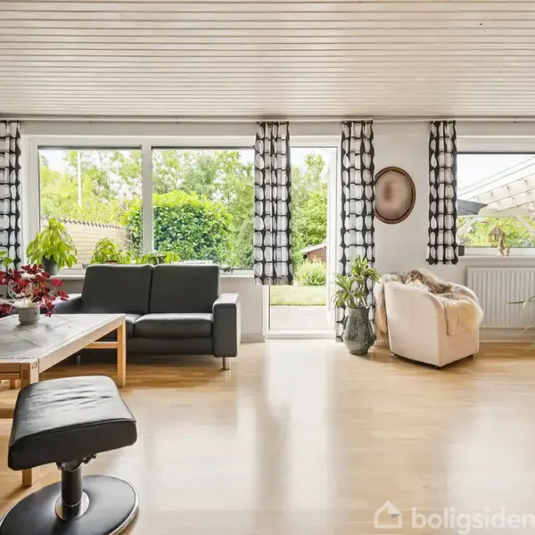 Stue med sorte lædermøbler og en beige lænestol. Store vinduer med sort-hvide gardiner giver udsigt til en have. Grønne planter står spredt rundt, og gulvet er af træ.